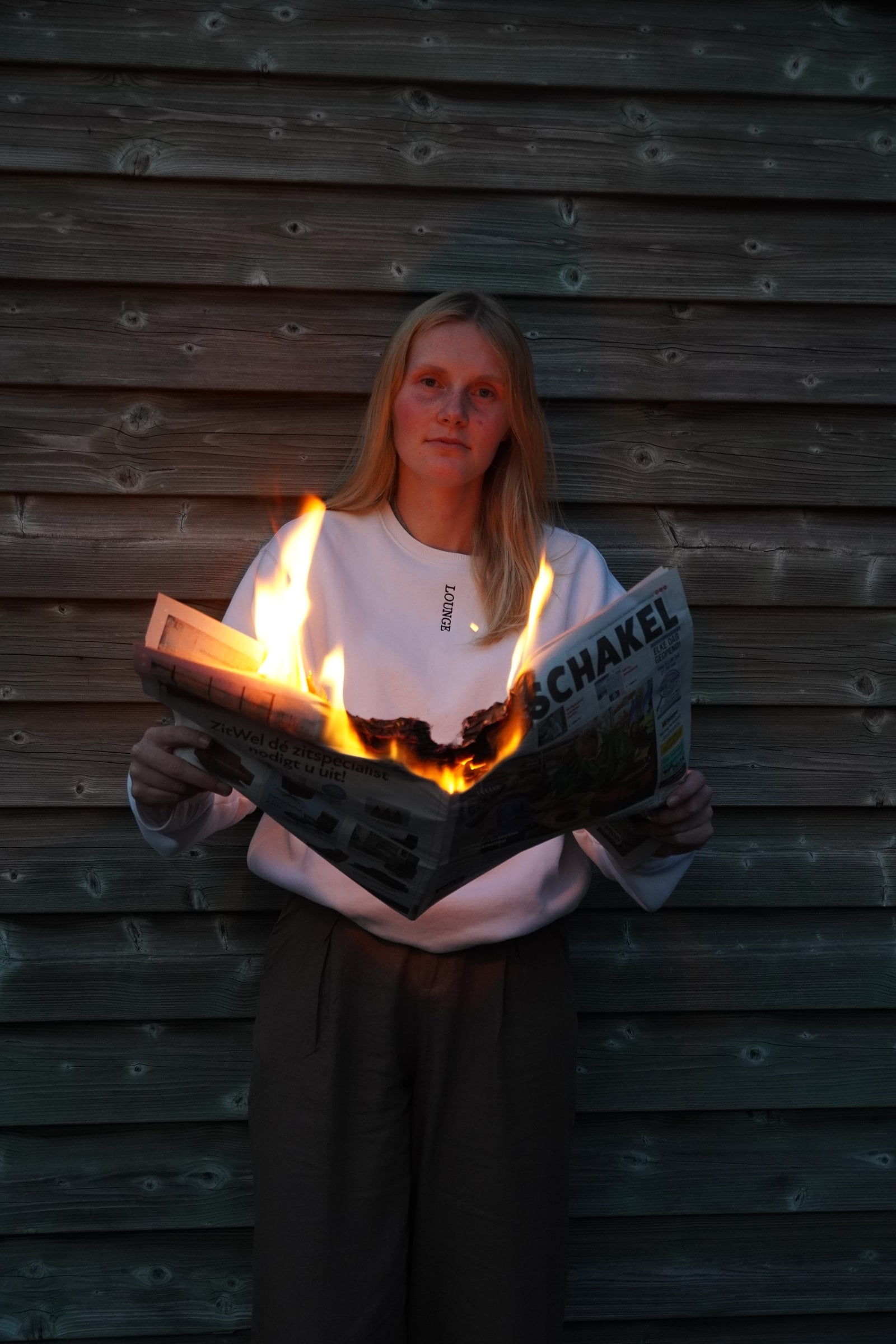 Persoon met lang blond haar en witte sweater staat voor een houten schuurwand en houdt een opengeslagen krant vast die in het midden in brand staat; het vuur verlicht de krant en de onderkant van de sweater oranje, terwijl de rest van het beeld in koel schemerlicht blijft (cover)