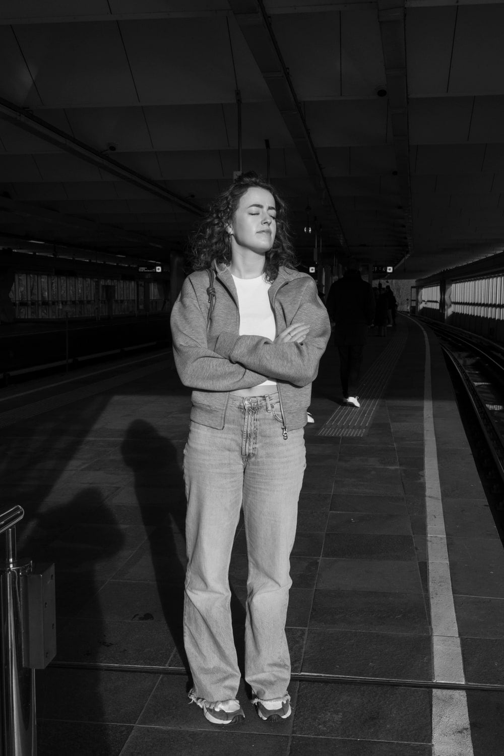 Vrouw met krullend haar staat met gesloten ogen en gekruiste armen in fel zonlicht op een overdekt metroperron; haar schaduw valt lang over de tegelvloer en achter haar loopt een donkere figuur weg over het perron.