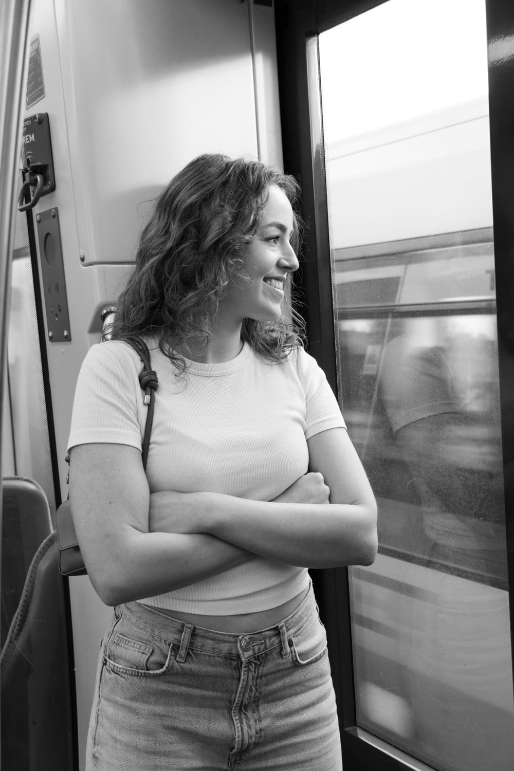 Lachende vrouw met lang krullend haar staat met gekruiste armen in een wit T-shirt en spijkerbroek naast de deur van een rijdende trein, met bewegingsonscherpte in het raam achter haar.