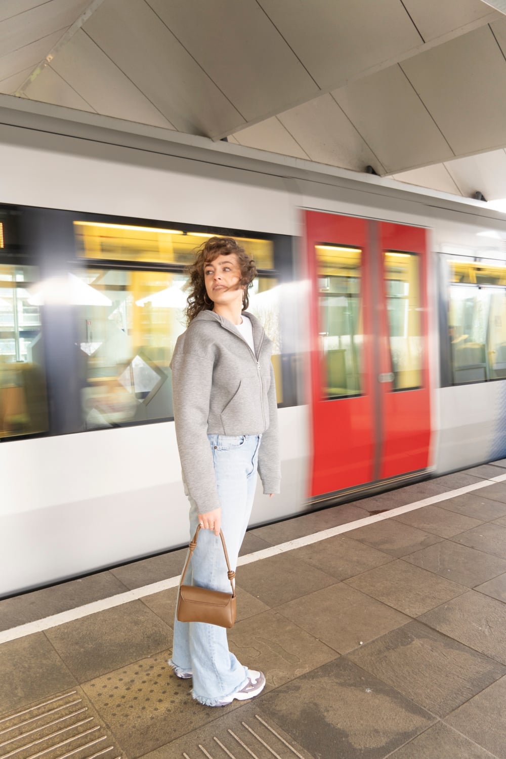 Vrouw in grijs wollen vest en wijde lichte jeans kijkt over haar schouder op een perron, terwijl achter haar een metro met rode deuren langsrijdt en tot beweging vervaagt.