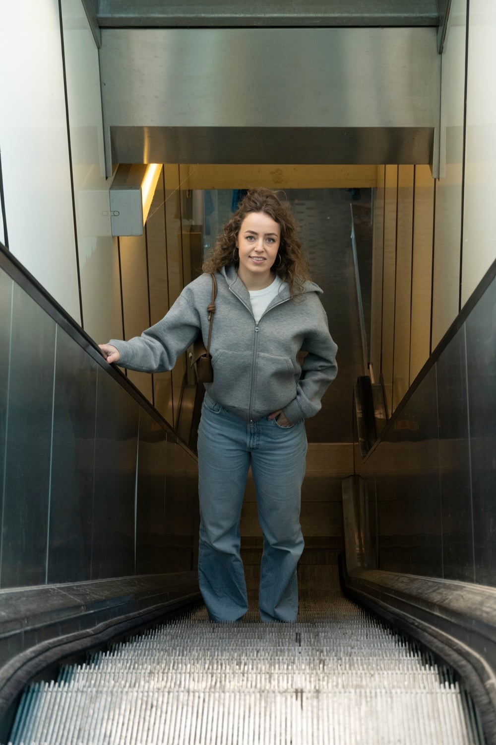 Vrouw staat frontaal bovenaan een roltrap, één hand aan de leuning en de andere in haar broekzak, en kijkt recht in de camera.