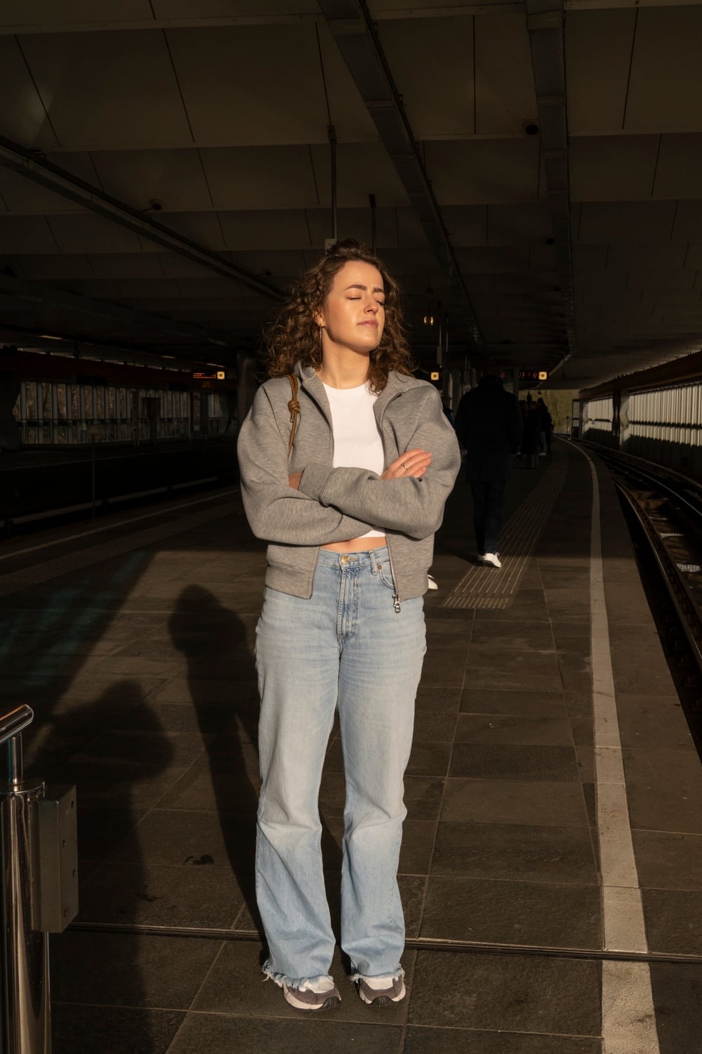 Vrouw staat met gesloten ogen en gekruiste armen in fel zonlicht op een overdekt perron; haar schaduw valt lang over de tegels.