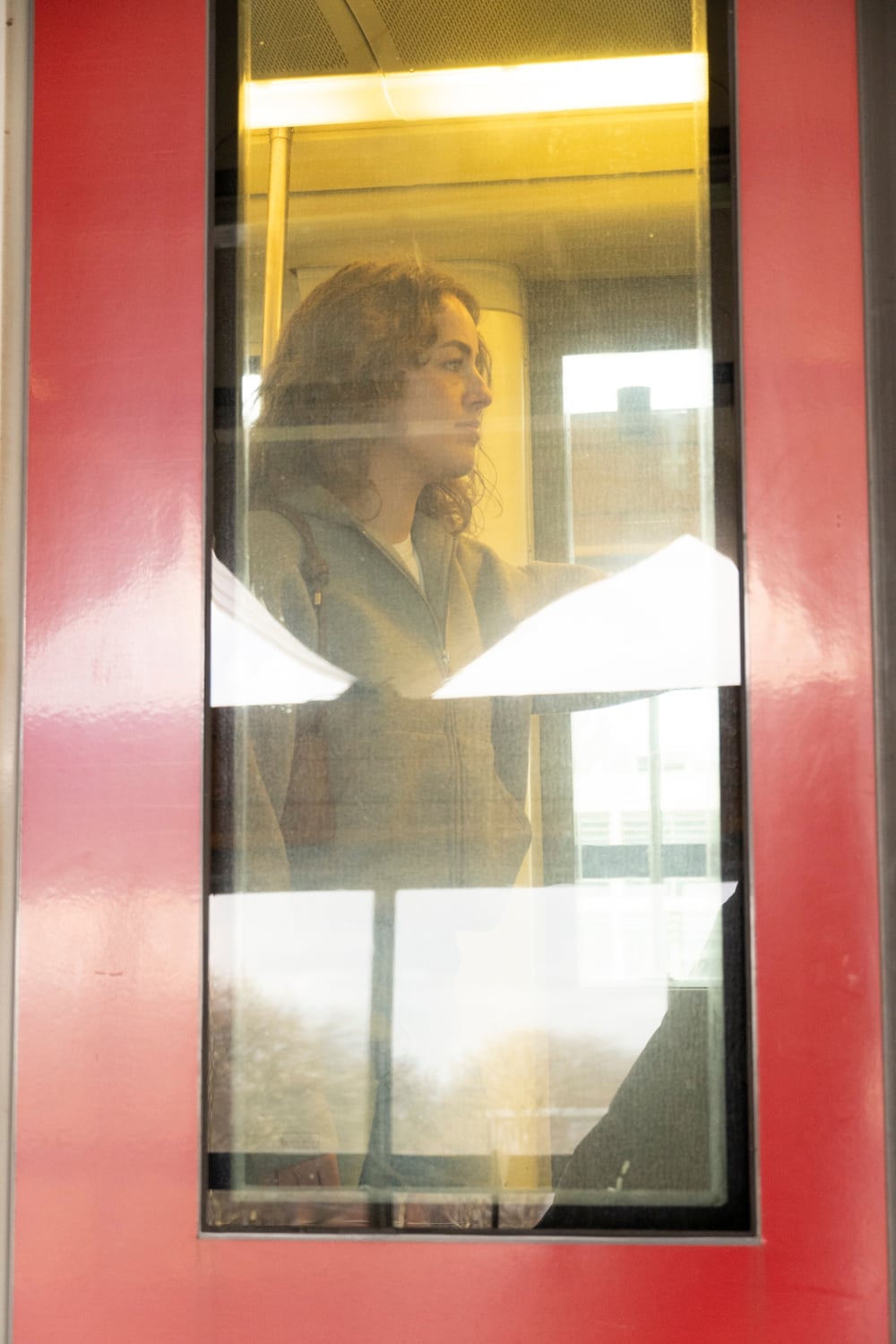 Vrouw in profiel achter het raam van een rode treindeur; in het glas weerspiegelen de gevels en bomen aan de overkant van het spoor.