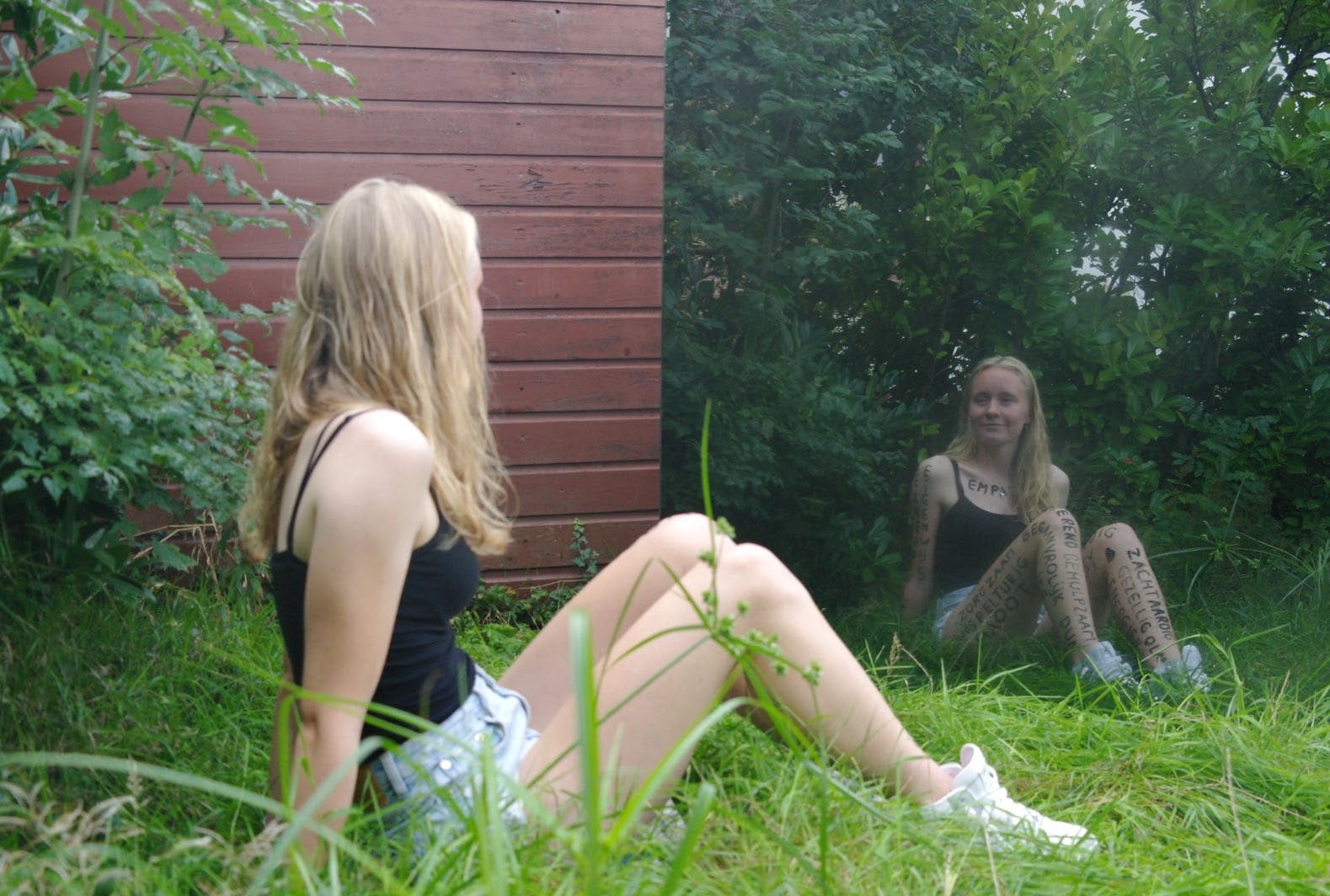 Vrouw met lang blond haar in een zwart hemdje en spijkershort zit in het hoge gras voor een grote spiegel die tegen een roodgeverfde houten schuurwand staat; in het spiegelbeeld kijkt ze glimlachend terug en zijn met stift geschreven woorden op haar armen, benen en borst te lezen
