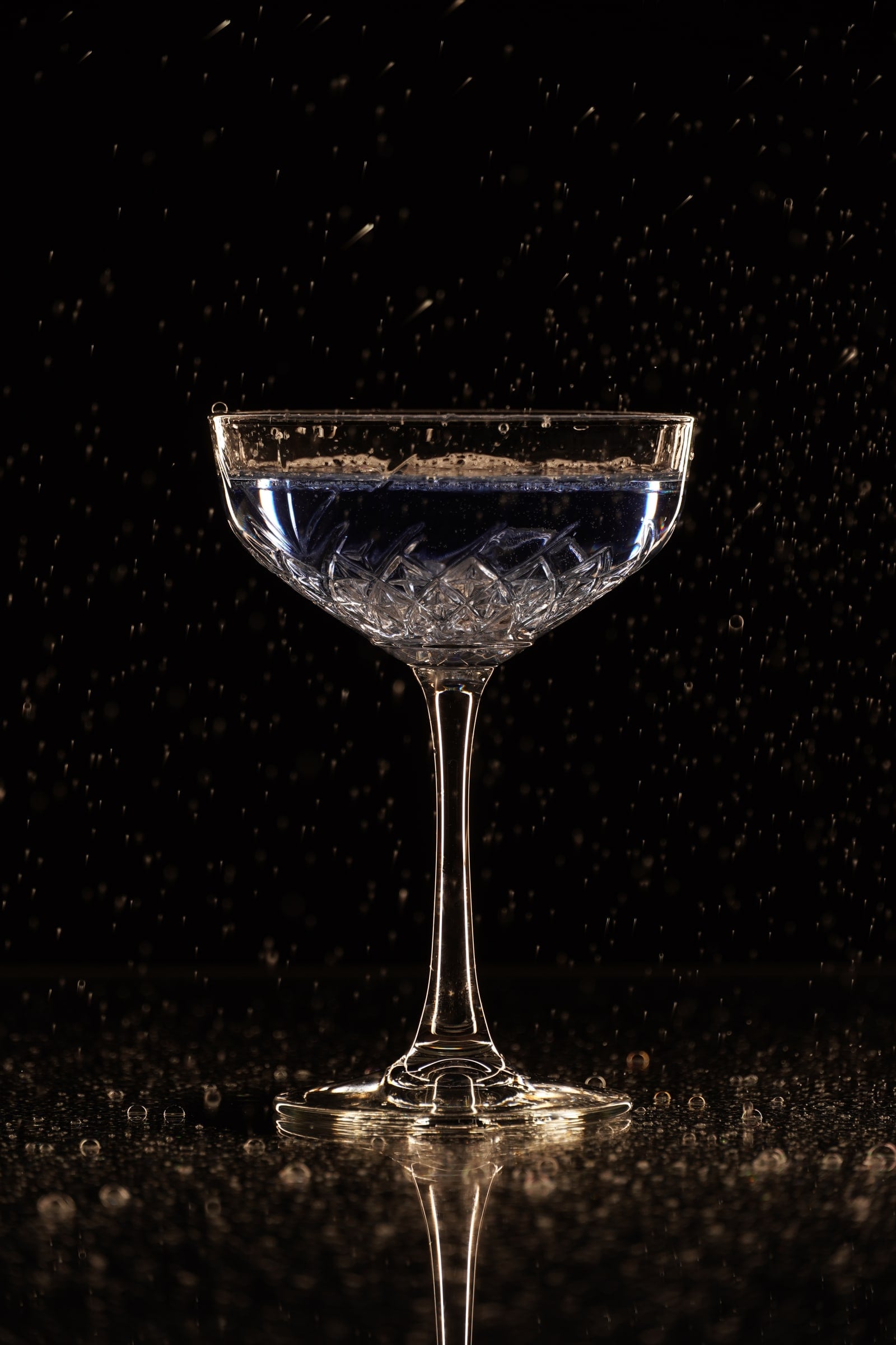 Geslepen coupeglas gevuld met blauwachtige vloeistof op een zwarte, natte ondergrond, met rondom bevroren waterdruppels in de lucht en warm randlicht langs de steel en de rand (cover)