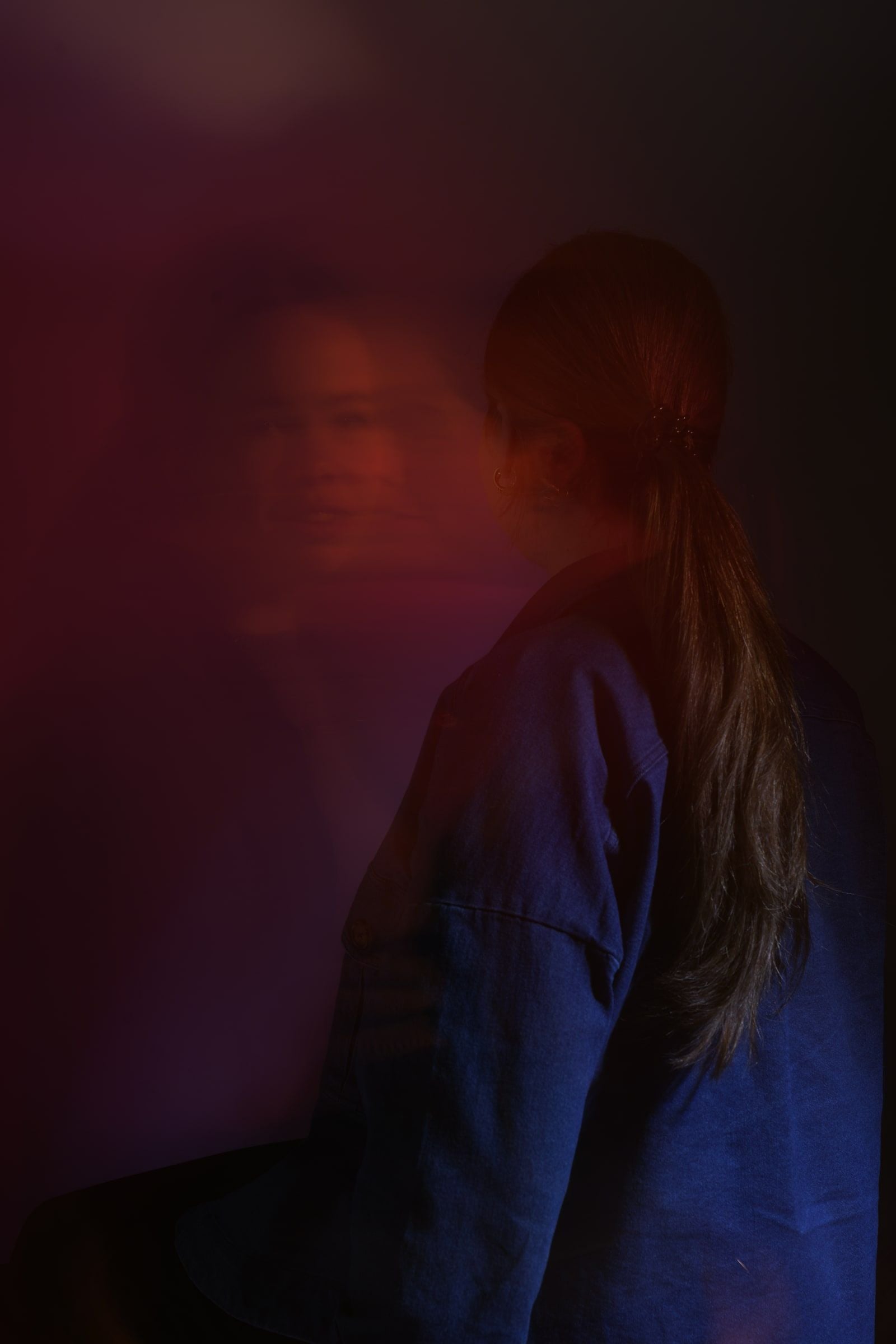 Vrouw met een lange staart in een spijkerjas gezien op de rug, met haar wazige spiegelbeeld in rood en paars licht naast haar