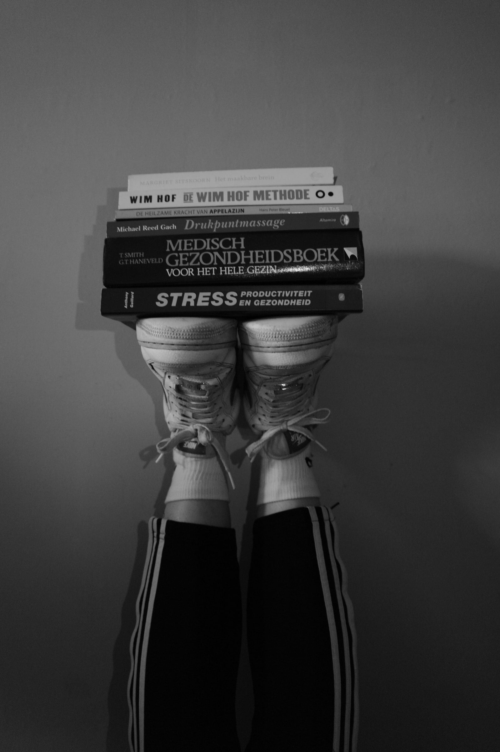 Zwart-wit opname van twee omhooggestoken benen in een sportbroek met streepjes, waarvan de sneakers een stapel boeken over gezondheid, stress en het brein op de neuzen balanceren