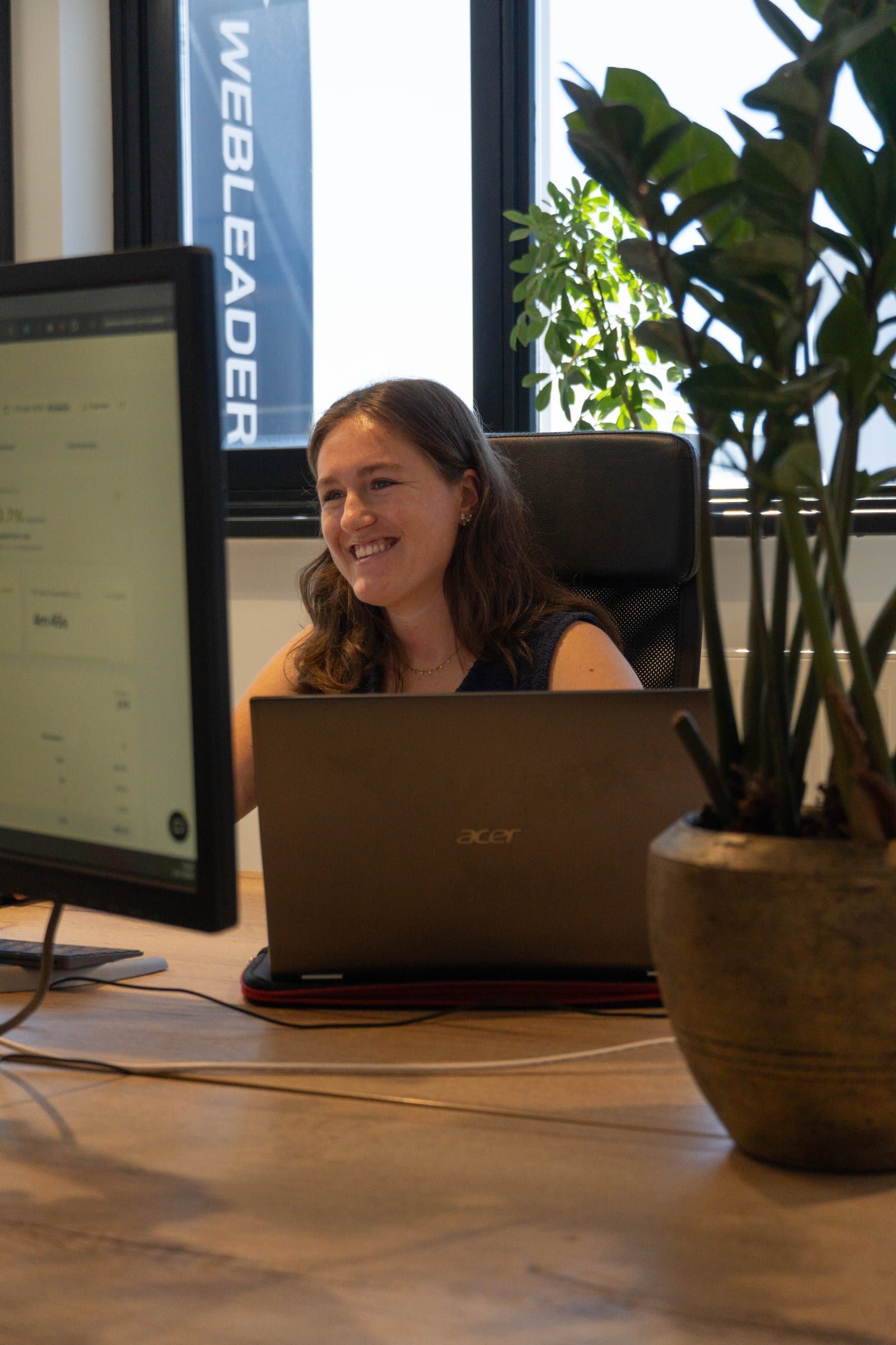 Lachende vrouw werkt achter een laptop en een groot scherm aan een houten bureau, met een Webleaders-banner en een grote plant achter haar.
