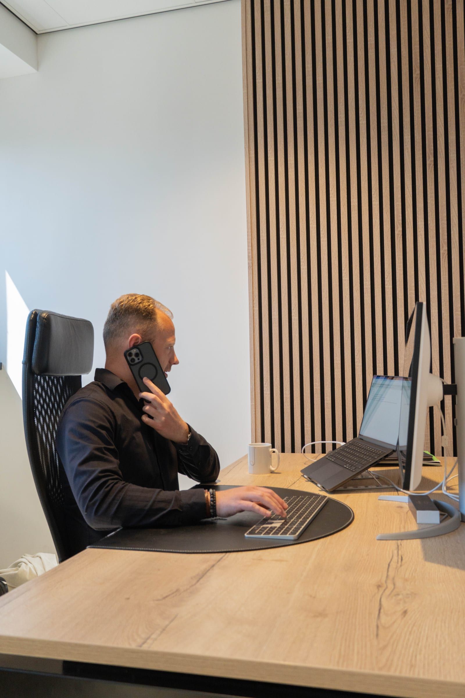 Man in zwart overhemd zit te bellen aan zijn bureau terwijl hij met zijn andere hand het toetsenbord bedient, tegen een wand van verticale houten latten.