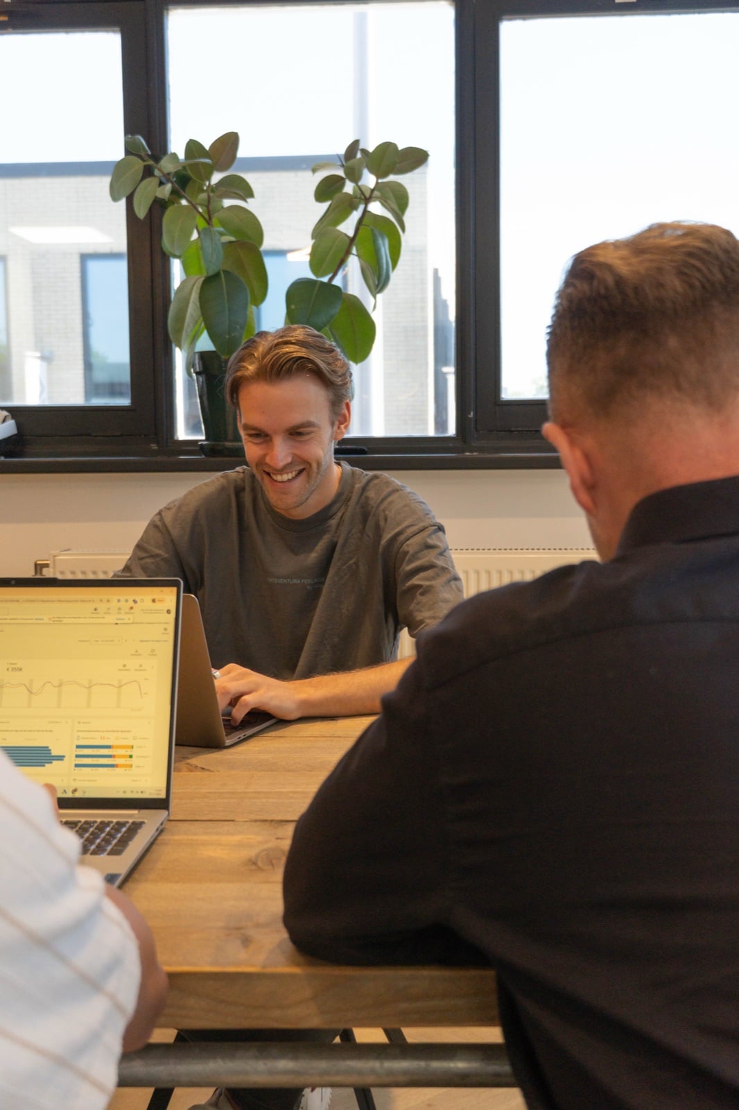 Lachende man werkt op een laptop aan een houten vergadertafel, met op de voorgrond twee collega's op de rug gezien.